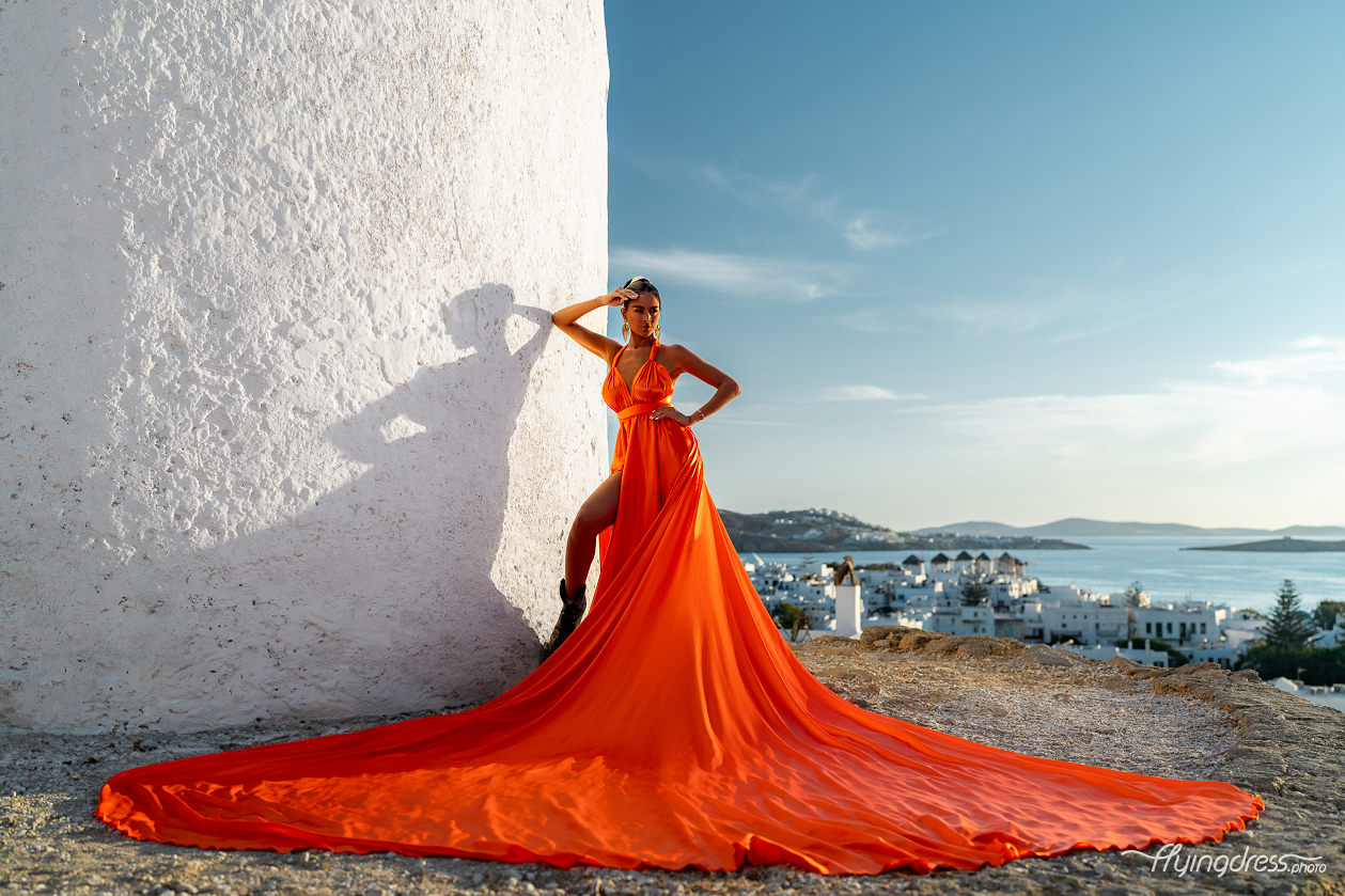 A woman in a striking orange gown poses confidently against a white-washed building in Mykonos, her dress flowing gracefully around her, with the beautiful Aegean Sea and white-washed buildings of the island in the distance.